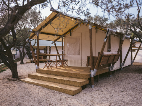 Tenda safari con terrazza in legno, tavolo e panca, circondata da alberi e ghiaia, ideale per glamping.