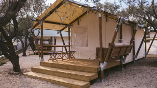 Tenda safari con terrazza in legno, tavolo e panca, circondata da alberi e ghiaia, ideale per glamping.