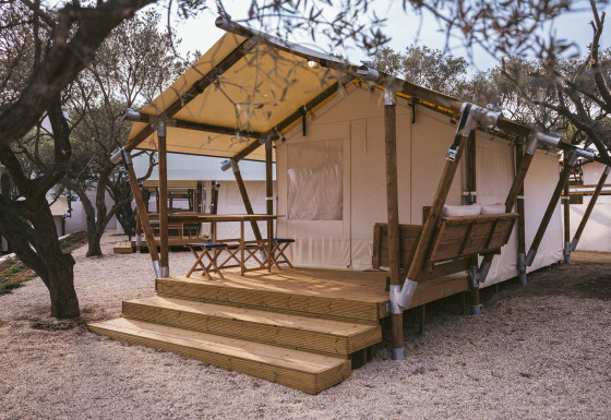 Tenda safari con terrazza in legno, tavolo e panca, circondata da alberi e ghiaia, ideale per glamping.