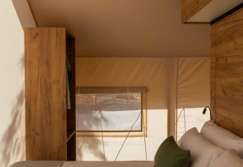 Interno di tenda safari con letto matrimoniale, cuscini verdi, aria condizionata e mobili in legno.