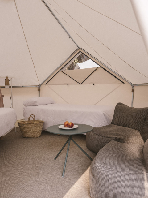 Interno della tenda safari Bell Tent Plus con due letti, poltrona, cesta e tavolo con frutta sul tappeto.