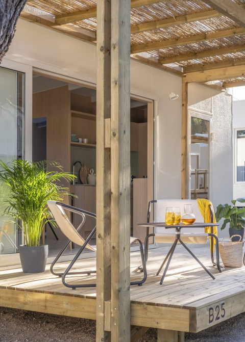 Véranda ensoleillée d'une tiny house avec terrasse en bois, deux chaises, une table et des plantes vertes.