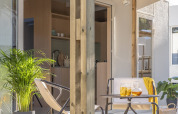 Porche soleado de una tiny house con terraza de madera, dos sillas, mesa con bebidas y plantas verdes.