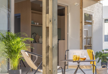 Zonnige tiny house veranda met houten terras, twee stoelen, tafeltje, drankjes en groene planten.