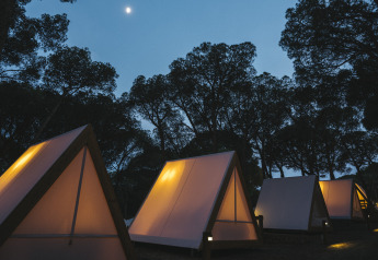 Tentes safari triangulaires éclairées au Wecamp Cala Montgó en Espagne, sous la lune, entourées d’arbres.