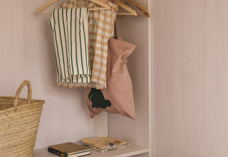 Minimalistische kast in een tiny house met hangende kleding, mand en boeken op de plank bij het bed.