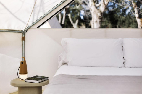 Inside a safari tent with a cozy bed, white pillows, and a nightstand holding a lamp and books.