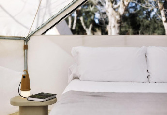 Innenansicht eines Safari-Zelts mit gemütlichem Bett, weißen Kissen und Nachttisch mit Lampe und Buch.