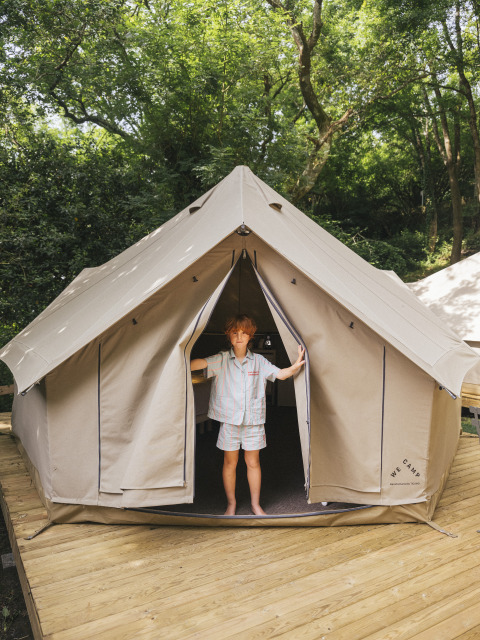 Un enfant se tient à l'entrée d'une tente safari Bell Tent sur une terrasse en bois dans une forêt verdoyante.