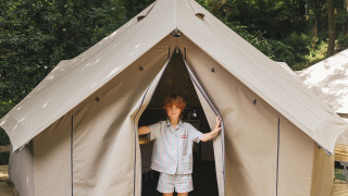 A child stands in the entrance of a safari Bell Tent on a wooden platform in a lush green forest.