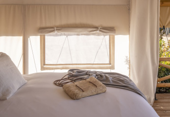 Letto accogliente nella tenda safari Glamping Couple, con plaid e borsa intrecciata vicino alla finestra.