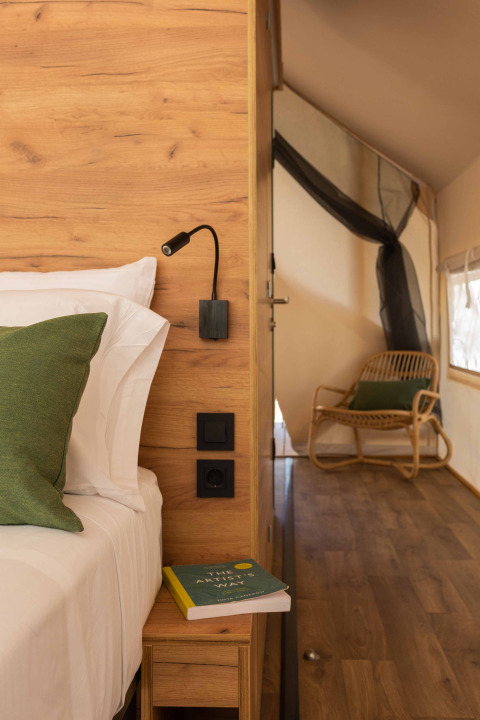 Vista interior de una tienda safari con paredes de madera, cama, cojín verde y silla al fondo.