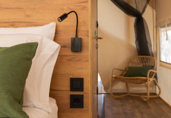 Vista interna di una tenda safari con pareti in legno, letto, cuscino verde e sedia sullo sfondo.