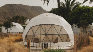Photo of a Domo dome with two chairs and a table outside, surrounded by palm trees and dry grass.