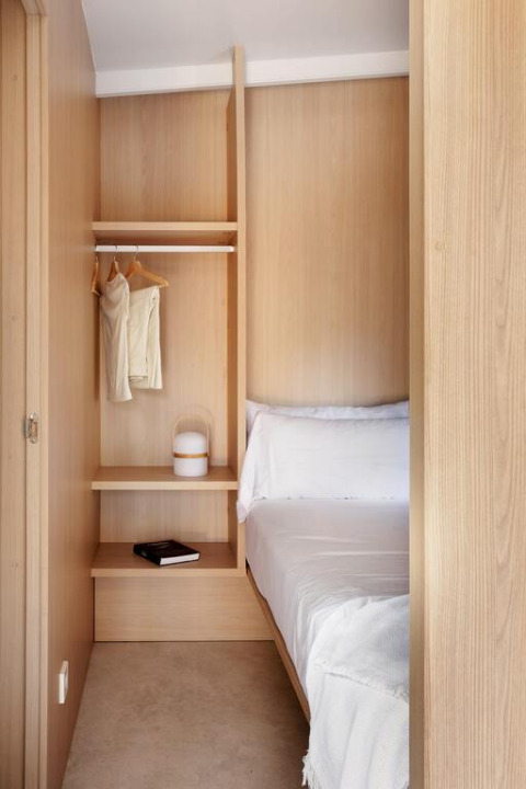 Camera luminosa e minimalista con armadio in legno, letto con biancheria bianca e libro su uno scaffale basso.