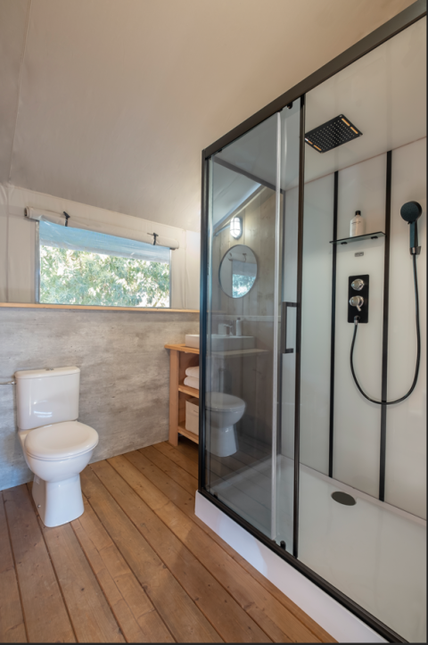 Moderne badkamer in een safaritent met houten vloer, glazen douche, toilet en raam met uitzicht.