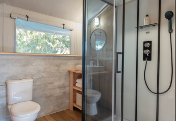 Bagno moderno in una tenda safari con pavimento in legno, box doccia in vetro, wc e finestra.