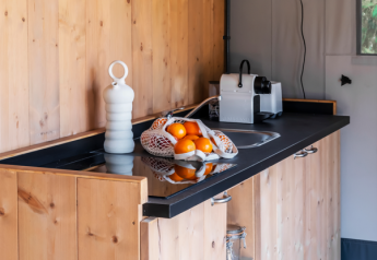 Cuisine de tente safari avec armoires en bois, plan de travail noir, cafetière et filet d’oranges fraîches.