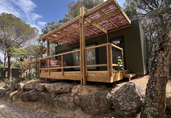 Mobil Home Family Plus lodge met houten veranda, omgeven door bomen en rotsen onder een blauwe lucht.