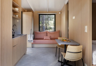 Interior de cabaña moderna con madera clara, cocina pequeña, mesa comedor y sofá frente a la ventana.