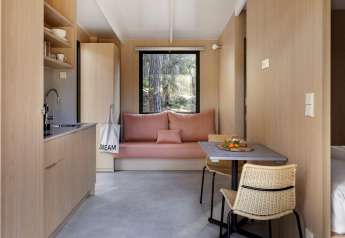 Intérieur de lodge moderne en bois clair avec kitchenette, coin repas et canapé face à une fenêtre sur la forêt.