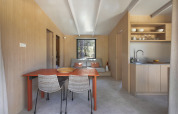 Moderne lichte lodge met eettafel, keuken en bank, omgeven door lichte houtpanelen en een groot raam.