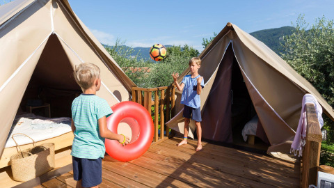 Twee kinderen spelen met een bal en een zwemband op een houten terras bij safaritenten in een groene omgeving.