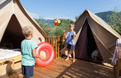 Two children play with a ball and float outside safari tents on a wooden deck surrounded by greenery.