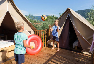 Twee kinderen spelen met een bal en een zwemband op een houten terras bij safaritenten in een groene omgeving.