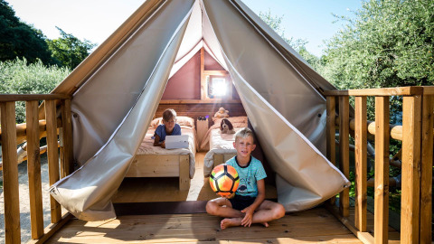 Niños juegan con un balón frente a una tienda safari con terraza y camas al aire libre en verano.