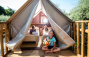 Two kids play with a soccer ball outside a safari tent with beds and a wooden deck on a sunny day.