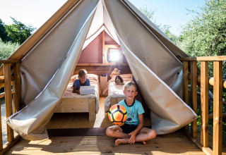 Kinderen spelen met een voetbal voor een safaritent met houten terras en bedden in een groene omgeving.