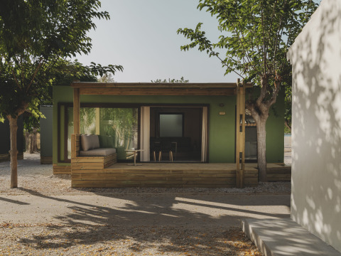 Groen MOBIL HOME FAMILY chalet met houten veranda en zitgedeelte, omringd door bomen in de zon