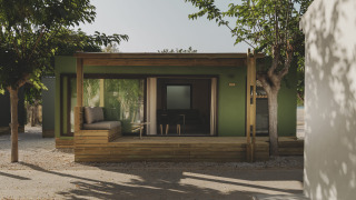 Groen MOBIL HOME FAMILY chalet met houten veranda en zitgedeelte, omringd door bomen in de zon
