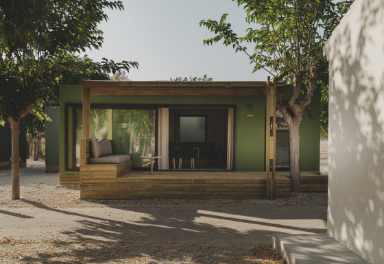 Groen MOBIL HOME FAMILY chalet met houten veranda en zitgedeelte, omringd door bomen in de zon