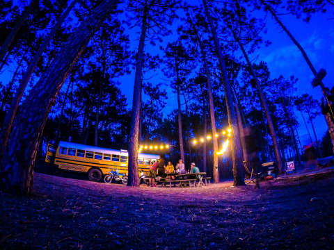 Scène de camping en forêt près d'une tiny house bus scolaire illuminée, Chianti Glamping Resort, Italie.