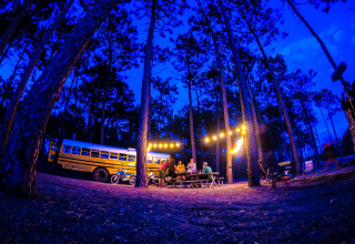 Scène de camping en forêt près d'une tiny house bus scolaire illuminée, Chianti Glamping Resort, Italie.