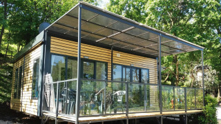 Next XL Panorama Lodge mit moderner Glasveranda, Holzfassade und umgeben von grünen Bäumen bei Sonnenschein.