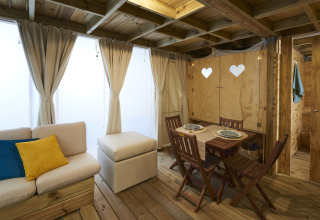 Woonruimte in Airlodge 2.0 met houten meubels, eethoek, bank met kussens en grote gordijnen voor ramen.