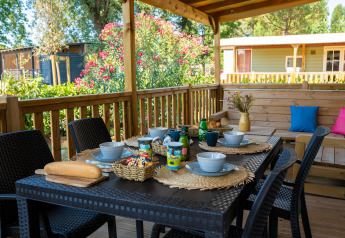Terraza al aire libre de una cabaña con mesa puesta, tazones, tazas, pan y fondo de vegetación.