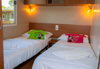 Chambre de lodge avec deux lits simples, t-shirts colorés, peluches, éclairage chaleureux et fenêtre.