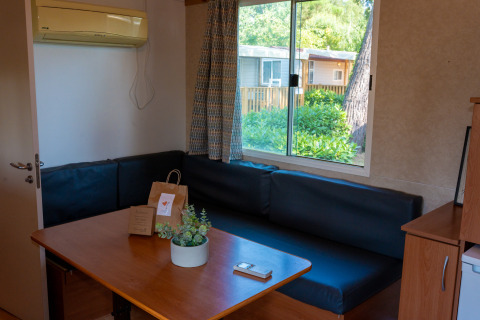 Interior de una cabaña con mesa de madera, sofá negro, ventana al jardín y aire acondicionado en la pared.