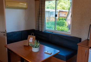 Intérieur de lodge avec table en bois, canapé noir en angle, vue sur jardin par la fenêtre et climatisation.