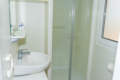 Baño pequeño y luminoso en una cabaña con lavabo, cabina de ducha, espejo, estante y ventana con papel higiénico.