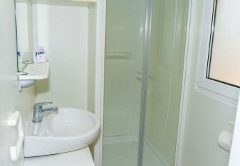 Baño pequeño y luminoso en una cabaña con lavabo, cabina de ducha, espejo, estante y ventana con papel higiénico.