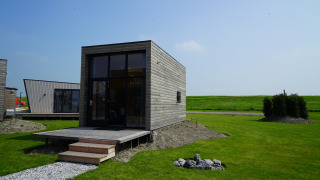 Modern Tiny House 2+2 with large windows and wooden siding set in a grassy landscape under blue sky.