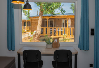 Vista desde el interior de una cabaña con escritorio y sillas hacia un porche de madera en Trasimeno Glamping Resort.