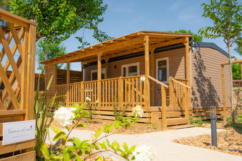 Moderna cabaña de madera Cottage Next XL Mercure con amplia veranda y vegetación en un día soleado.