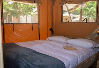 Interno di una tenda safari con un comodo letto matrimoniale, cuscini, lampade e finestre sulla natura.