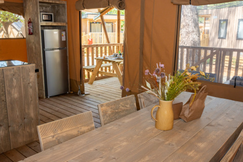 Interieur van een safaritent met houten tafel, koelkast, bloemenvaas en uitzicht op het terras.